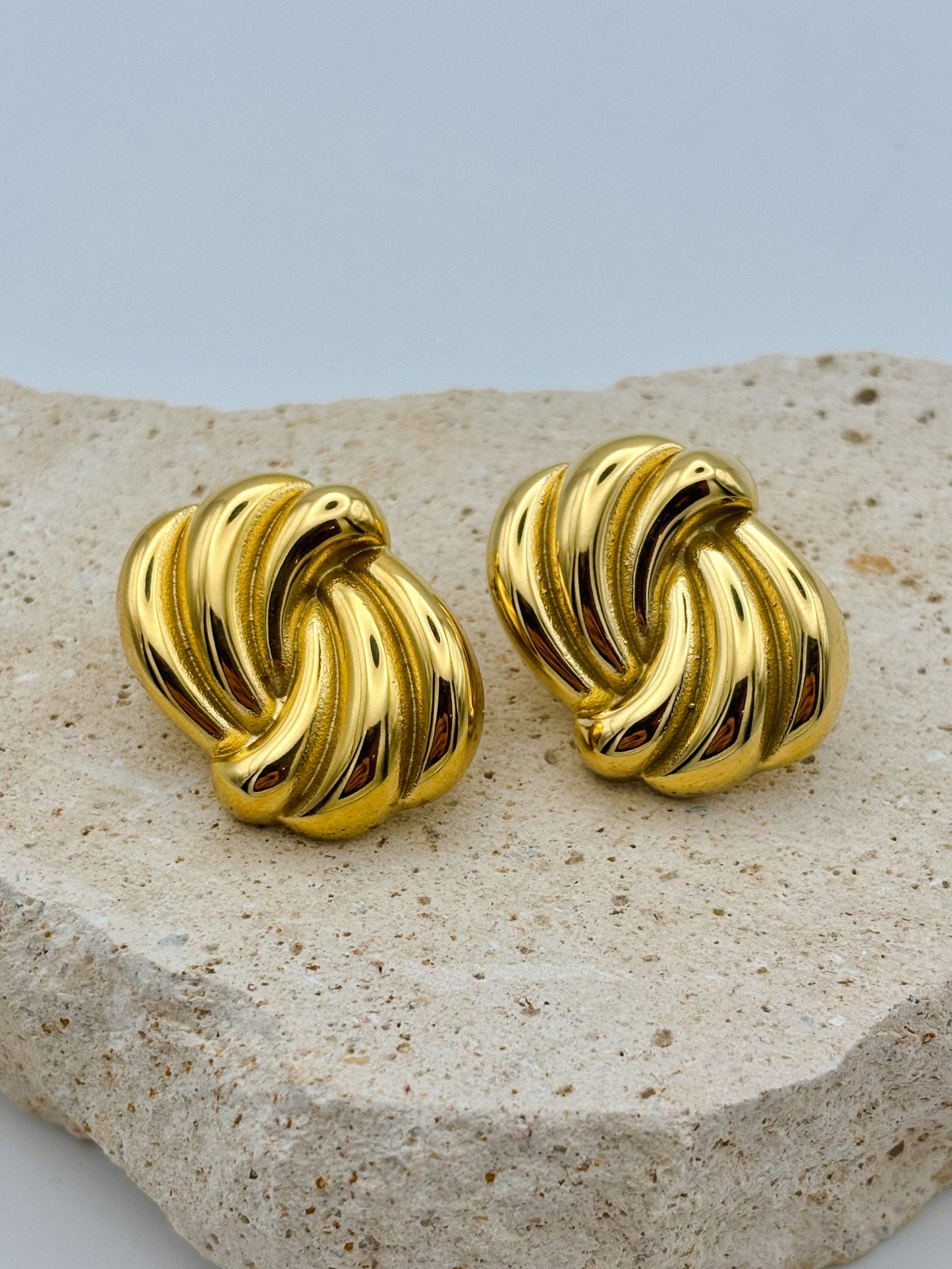 Pair of vintage-style gold-tone clip-on earrings with a twisted, sculptural design, displayed on a textured beige stone surface. Retro. Vintage. Chunky earrings.