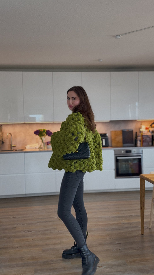 A woman wearing a chunky, textured olive green crochet flower jacket, dark gray leggings, and black boots, standing in a modern kitchen with white cabinets and wood floors.