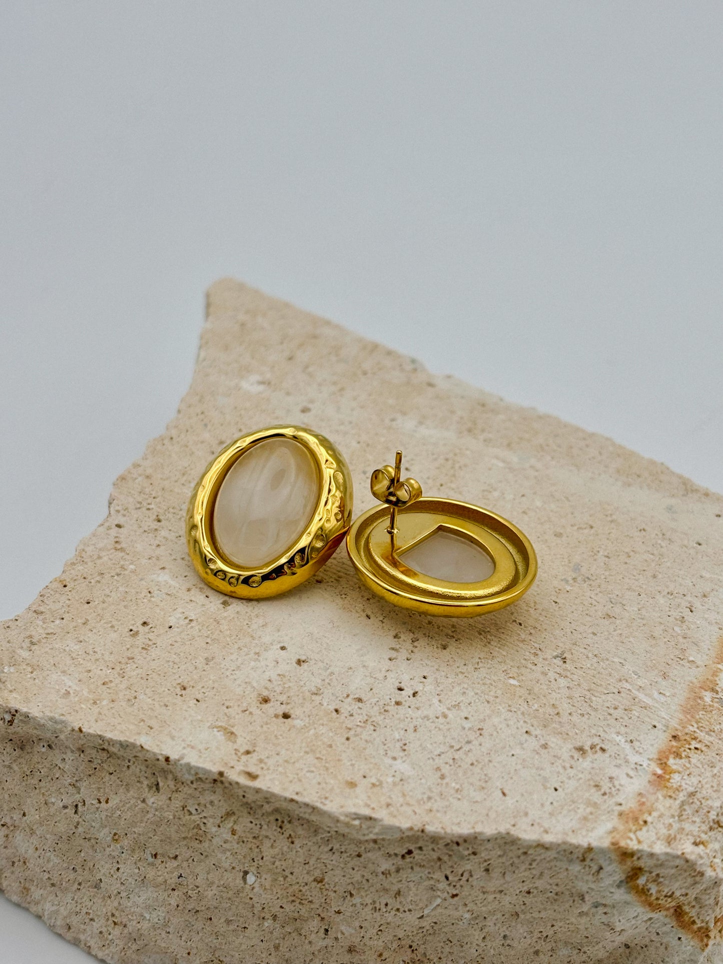 Paar goldene Ohrringe mit ovalem, cremefarbenem Stein – einer zeigt die Vorderseite, der andere die Rückseite mit Stecker – arrangiert auf hellem Naturstein
