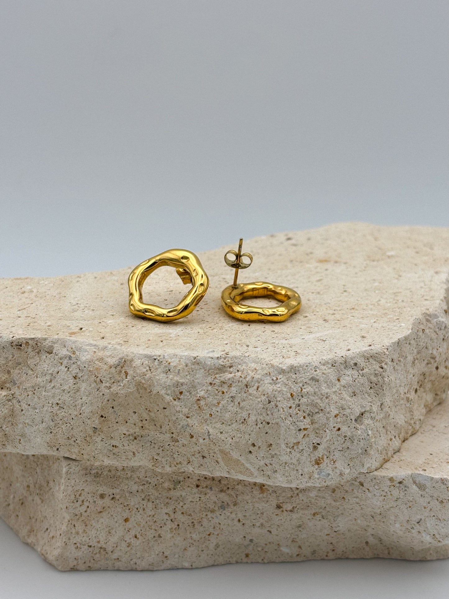 Organic-shaped gold stud earrings with textured open circle design, displayed on layered natural stone — modern minimalist jewelry with artistic flair