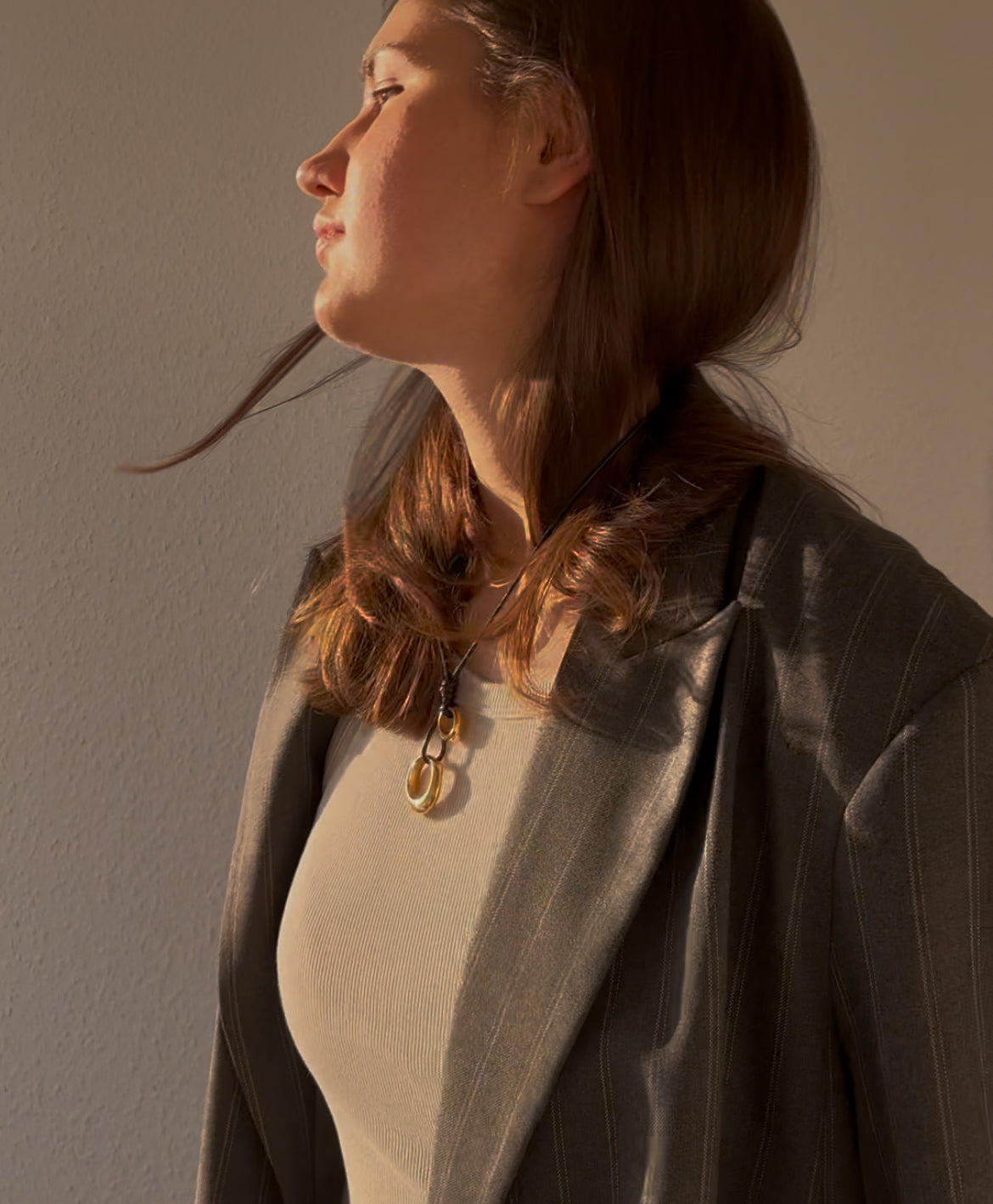 Woman wearing layered stainless steel pendant necklace on black cord, styled with neutral tank top and oversized pinstripe blazer — modern minimalist jewelry with artistic flair in warm natural light