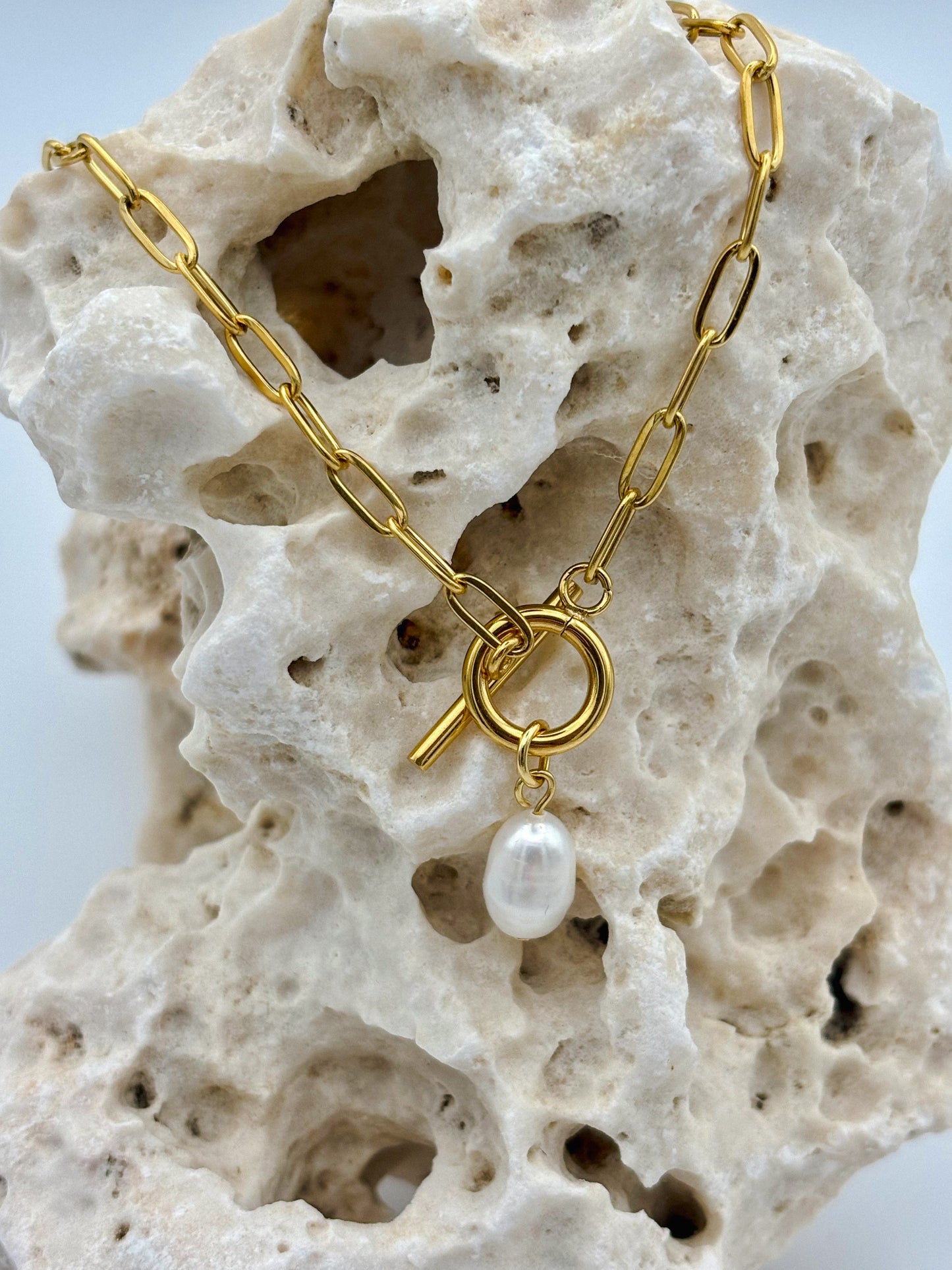 Gold paperclip chain necklace with toggle clasp and dangling freshwater pearl pendant, draped over porous natural stone — modern minimalist jewelry with organic elegance