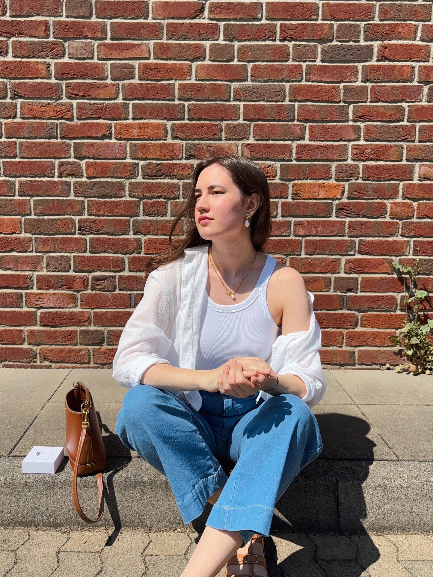 a woman is sitting on a floor in front of a brick wall in white tank top, white blouse, blue jeans and brown bag, under the direct sunlight wearing pearl necklace and earrings
