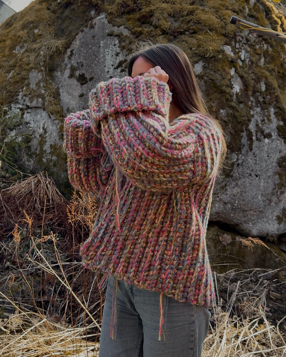 Person wearing a chunky knit sweater in a natural setting with rocks and dry grass.