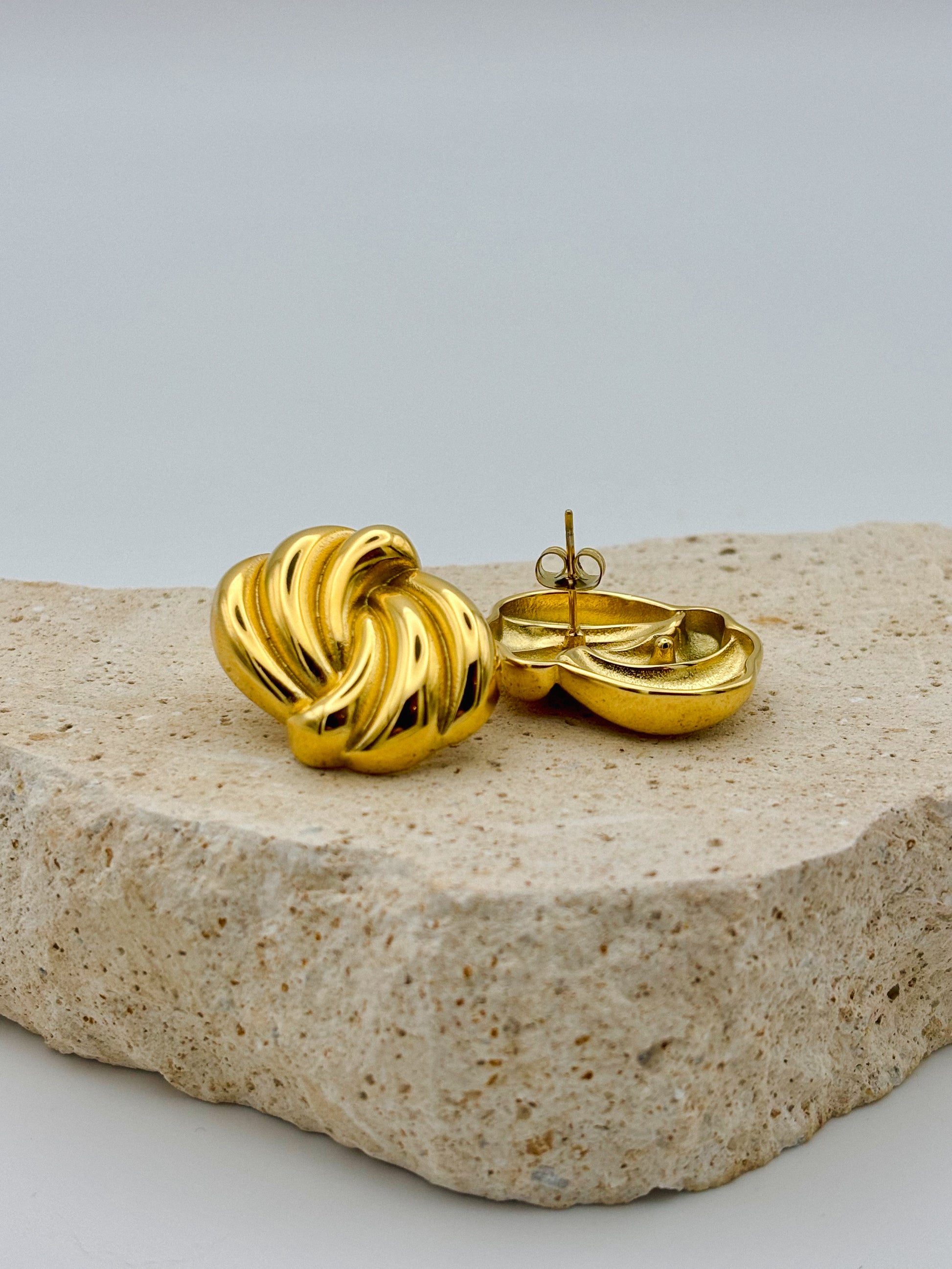 Gold-tone twisted earrings with a sculptural, ridged design. One earring is shown facing forward, while the other is laid on its back to reveal the post and clip-on backing, both resting on a rough beige stone surface. Woman wearing sculptural gold earring in sunlight with shadow play.  Retro. Vintage. Chunky earrings. Hand-made.