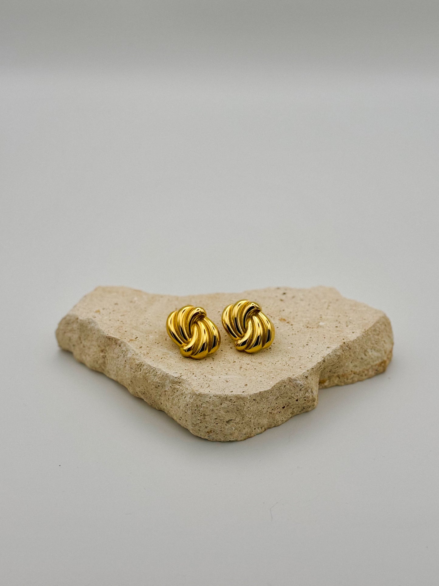 Pair of vintage-style gold-tone clip-on earrings with a twisted, sculptural design, displayed on a textured beige stone surface.Woman wearing sculptural gold earring in sunlight with shadow play.  Retro. Vintage. Chunky earrings. Hand-made. Hand made jewellery.