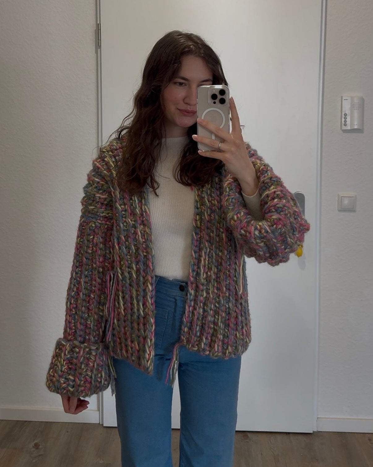 Person taking a mirror selfie wearing a patterned cardigan, white shirt, and blue jeans in a room with a white wall.