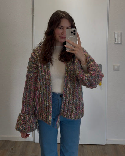Person taking a mirror selfie wearing a patterned cardigan, white shirt, and blue jeans in a room with a white wall.