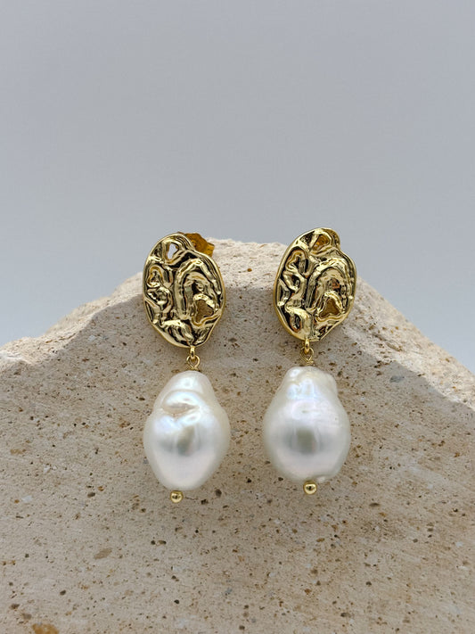 Goldene Ohrringe mit organisch geformtem Stecker und großer, barockähnlicher Perle als Anhänger, arrangiert auf hellem Naturstein.