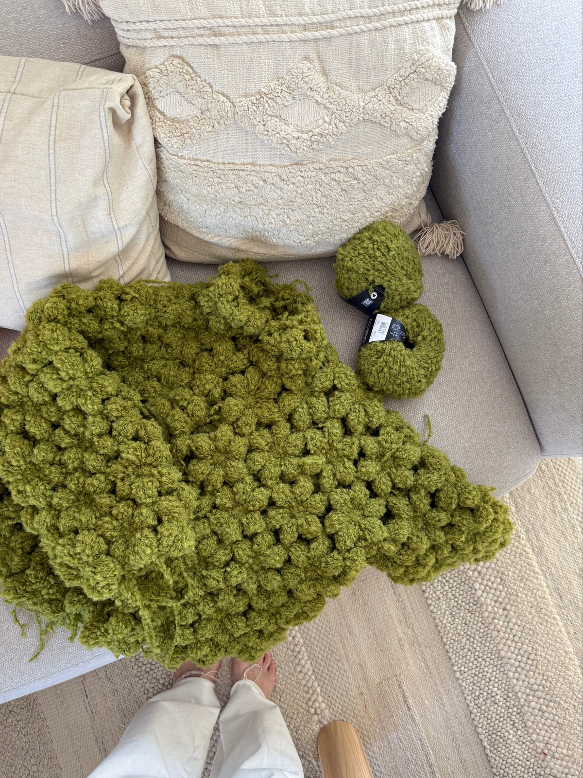An olive green chunky crochet jacket is draped over a light gray sofa, with matching green boucle yarn beside it. A person's legs in white pants are visible at the bottom of the frame, and the setting is a cozy living room with neutral-toned pillows and a woven rug.