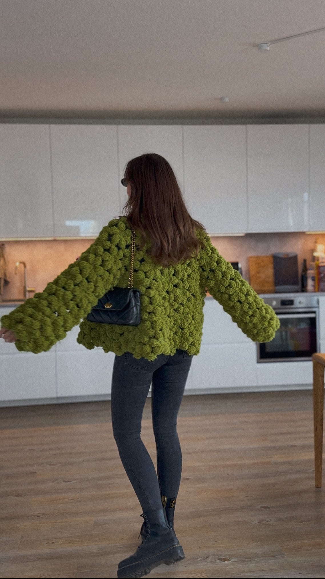 A woman wearing a chunky olive green flower crochet sweater, dark gray leggings, and black boots, standing in a modern kitchen with white cabinets.