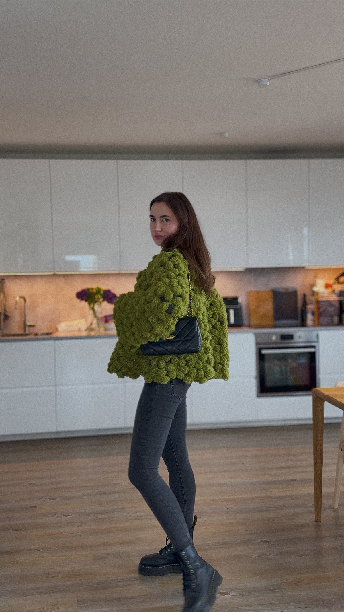 A woman wearing a chunky, textured olive green crochet flower jacket, dark gray leggings, and black boots, standing in a modern kitchen with white cabinets and wood floors.