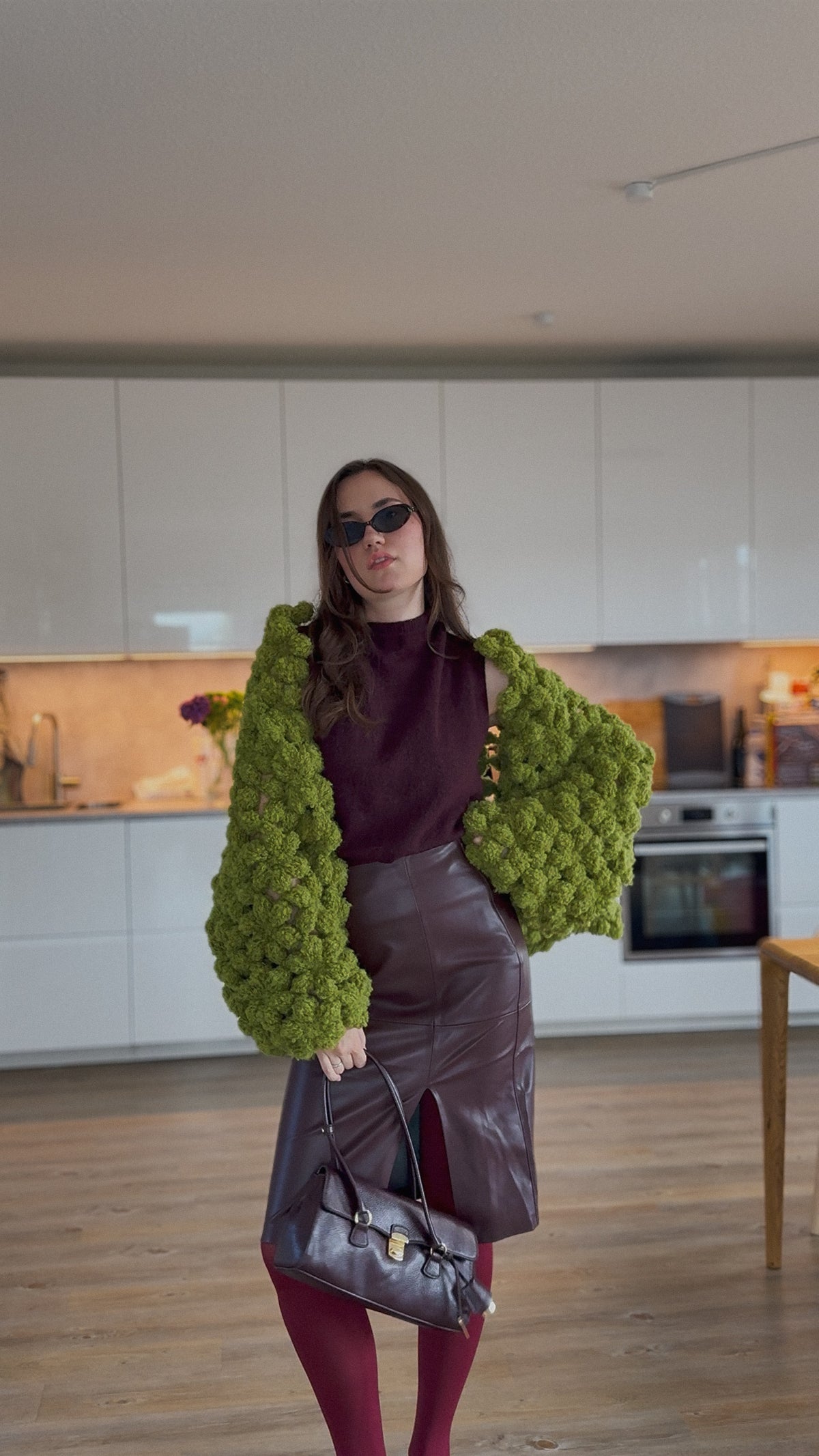 A woman stands in a modern kitchen wearing a chunky lime green boucle flower jacket over a dark plum top, a dark brown faux-leather skirt, burgundy tights, and holding a dark brown handbag.