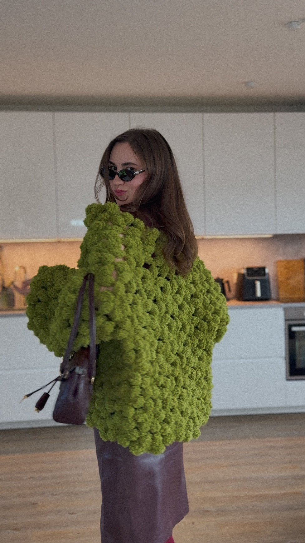 A woman wearing a chunky, oversized lime green textured crochet flower sweater and a dark brown leather skirt, holding a dark brown handbag in a modern kitchen setting.