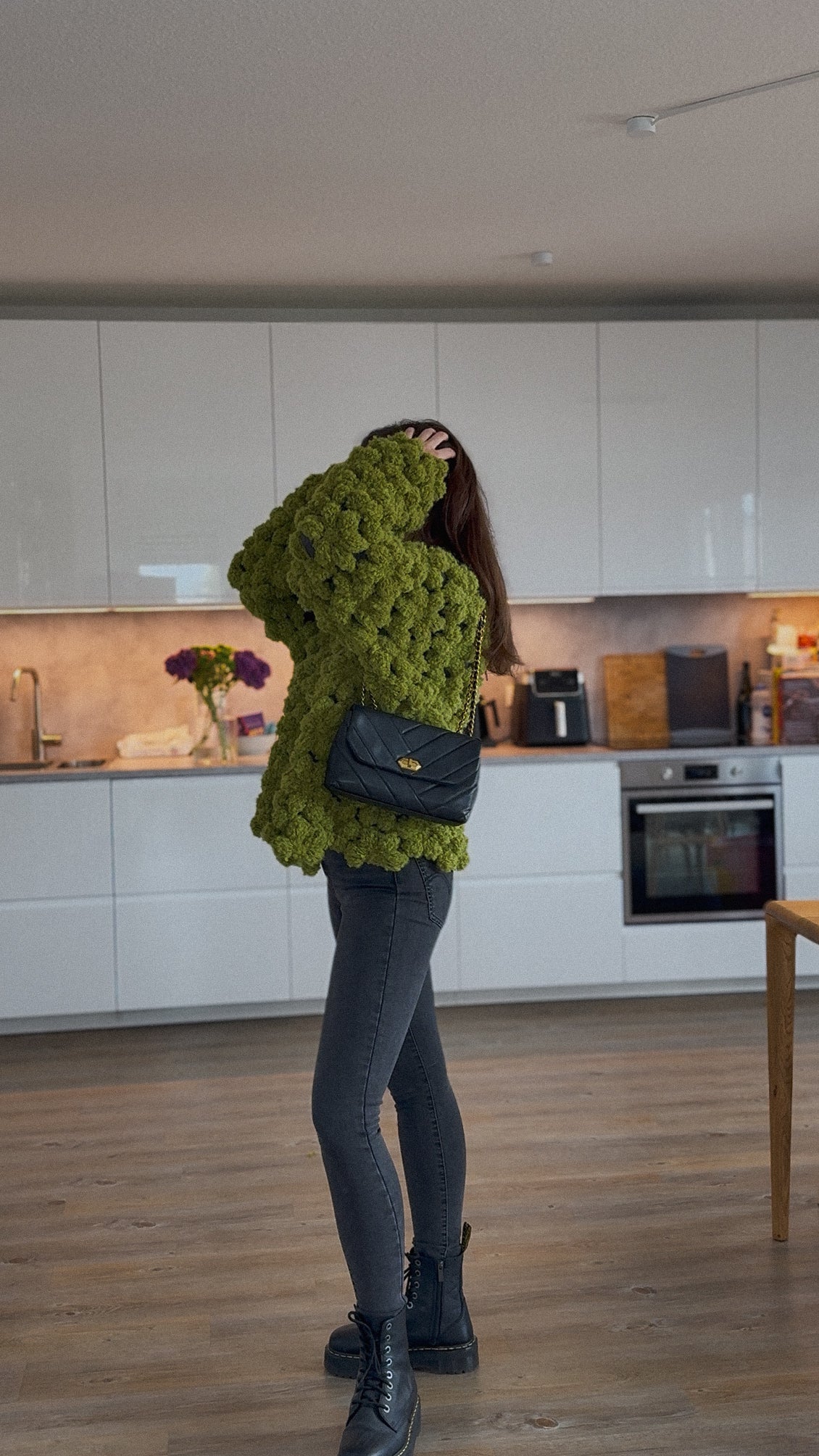 A woman stands in a modern kitchen wearing a chunky, textured olive green flower boucle sweater, dark gray skinny jeans, black boots, and carrying a black quilted shoulder bag.