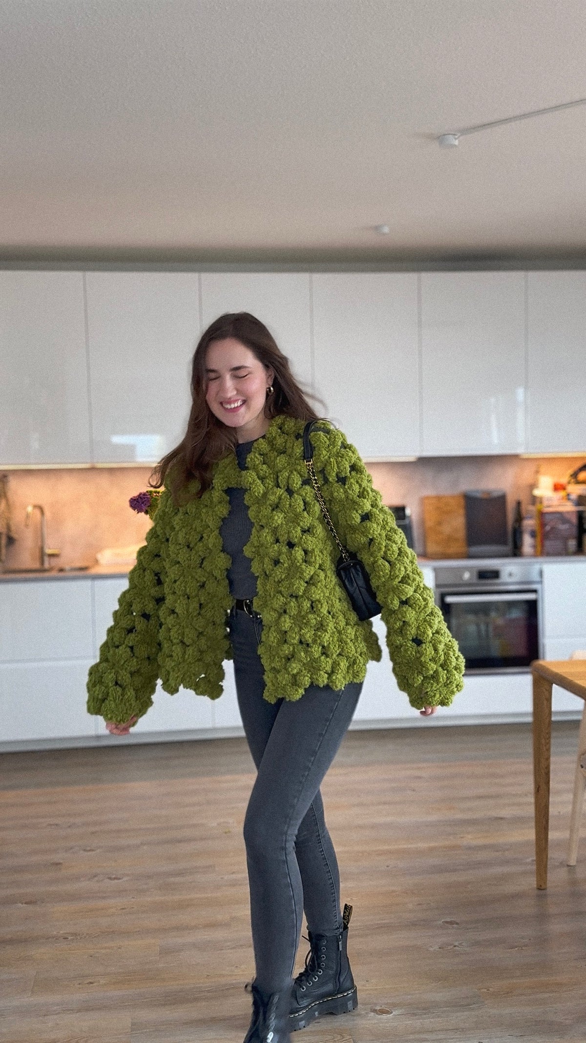 A woman wearing a chunky olive green boucle crochet flower jacket, dark gray skinny jeans, and black boots, standing in a modern kitchen with white cabinets and wood floors.