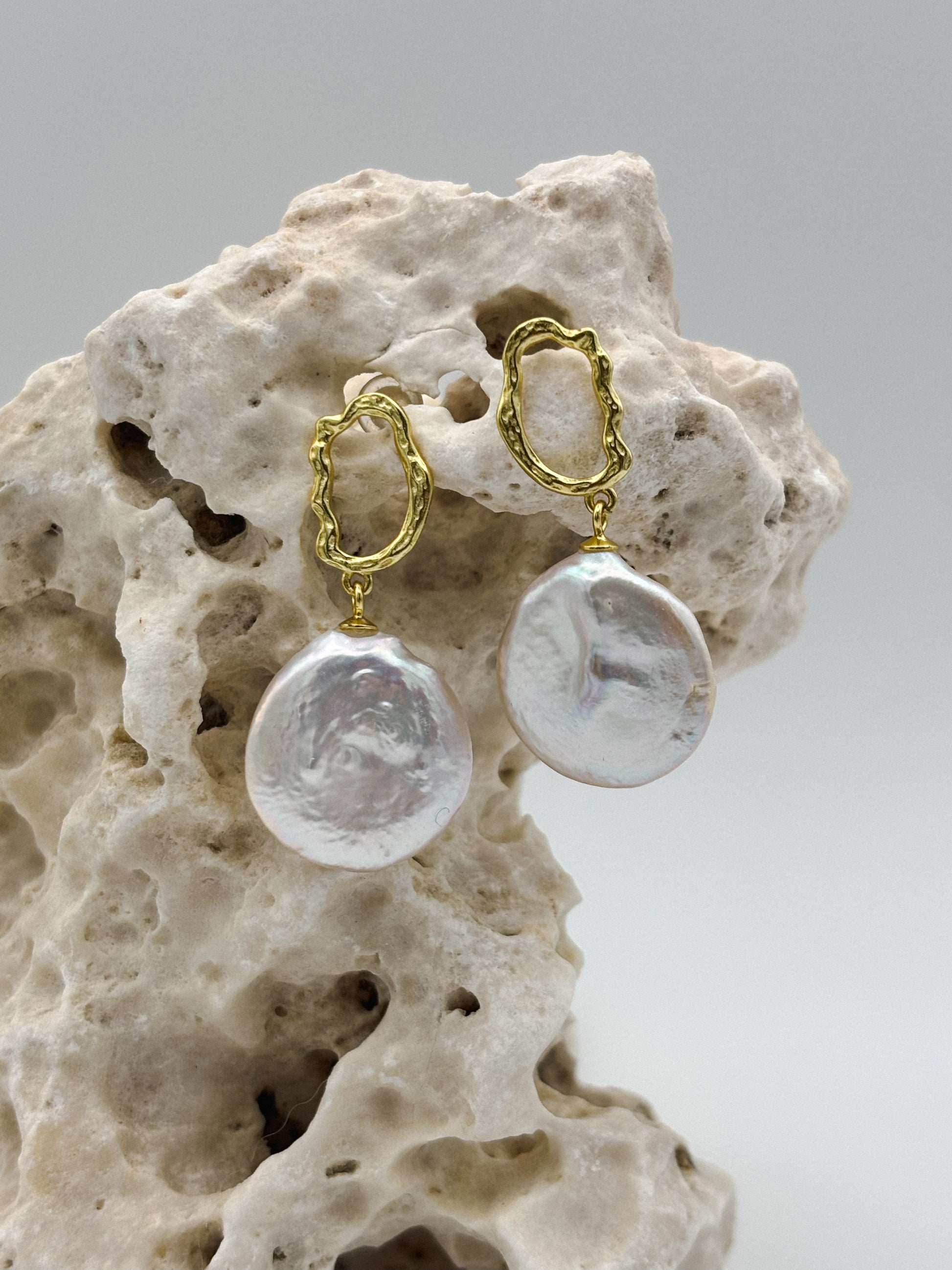Gold drop earrings with irregular baroque freshwater pearls, hanging from textured oval studs, displayed on natural porous stone — elegant handcrafted jewelry with organic charm