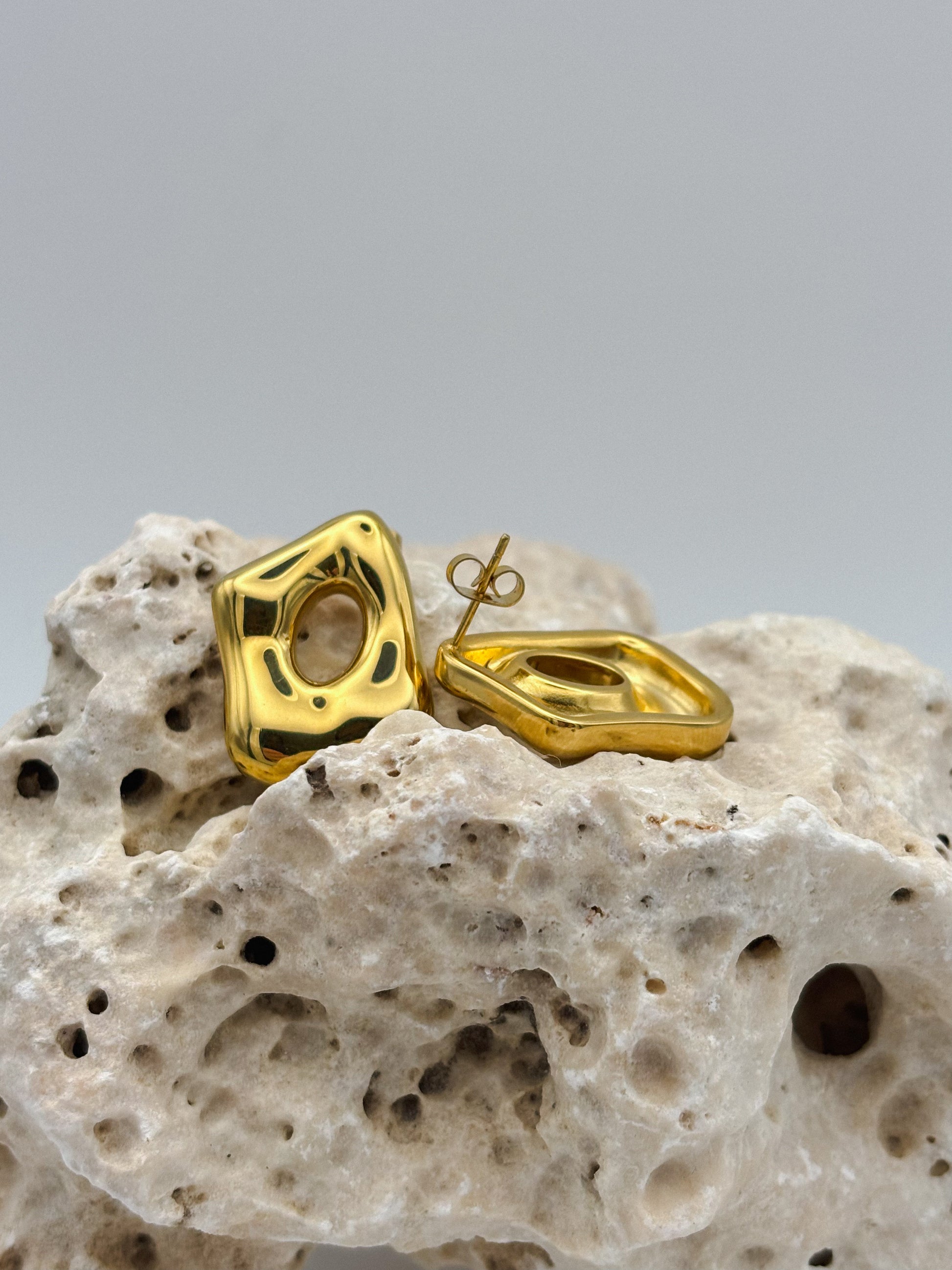 Modern gold stud earrings with abstract square shape and organic hollow center, displayed on textured stone — bold minimalist design for contemporary jewelry lovers