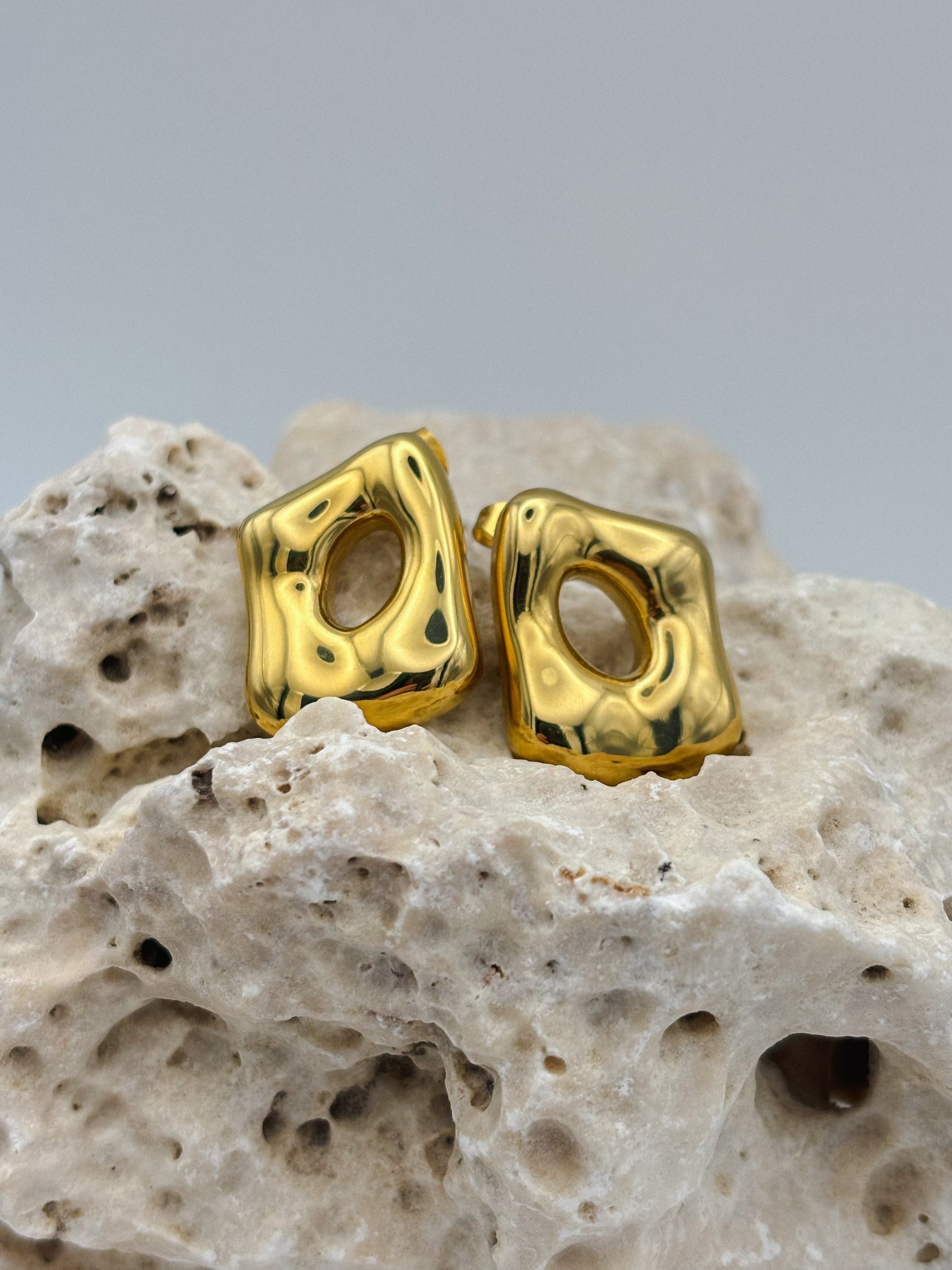 Modern gold stud earrings with abstract square shape and organic hollow center, displayed on textured stone — bold minimalist design for contemporary jewelry lovers