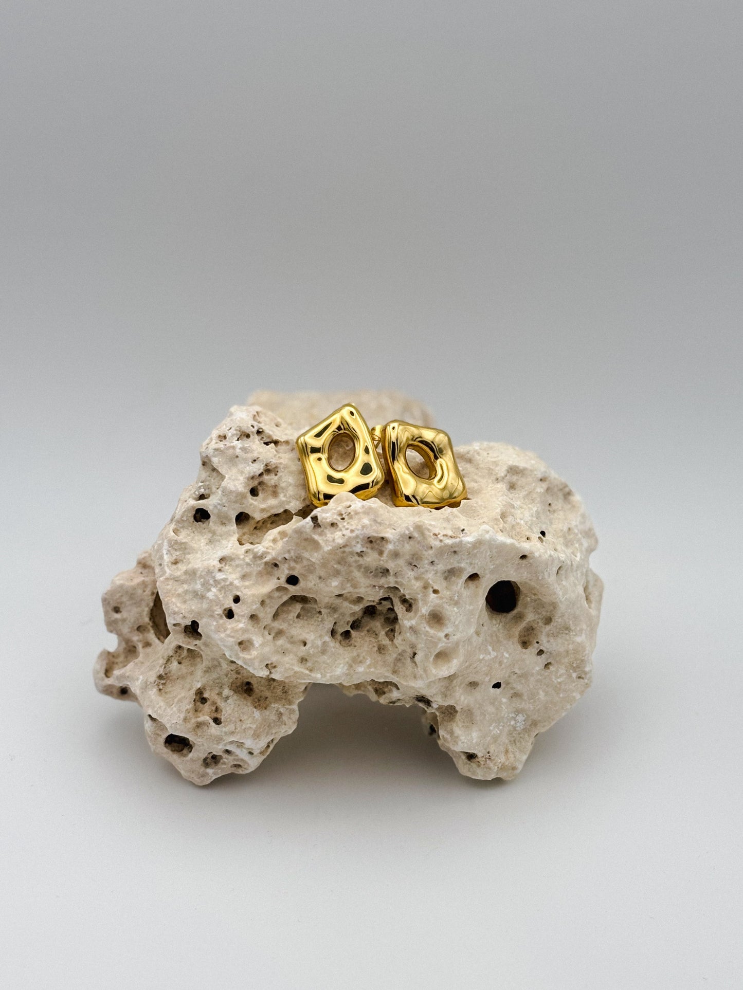 Gold abstract square-shaped stud earrings with organic cutout design, displayed on natural porous stone — modern artistic jewelry with minimalist flair