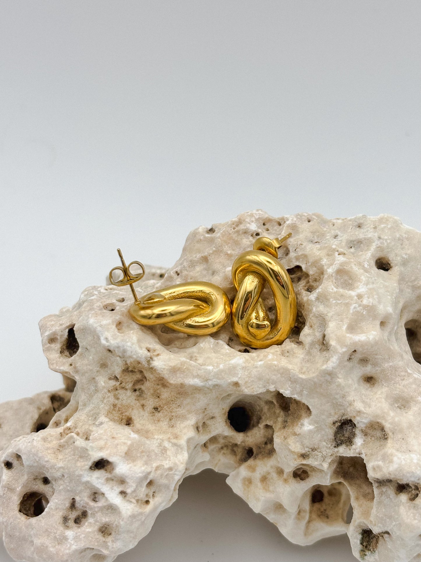 Gold abstract knot stud earrings with sculptural twist design, displayed on porous natural stone — modern artistic jewelry with a minimalist edge