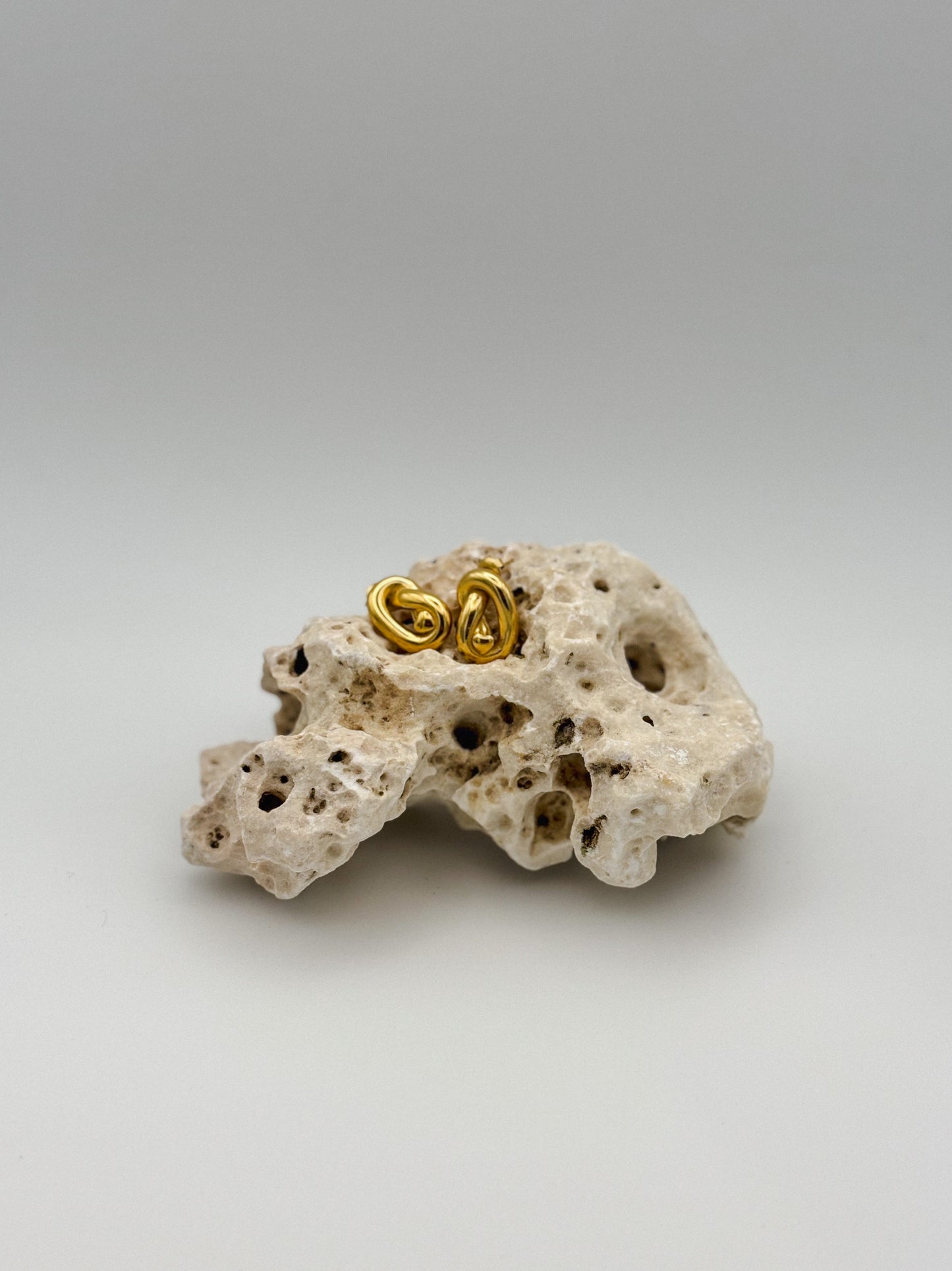 Gold abstract knot stud earrings with sculptural twist design, displayed on porous natural stone — modern artistic jewelry with a minimalist edge