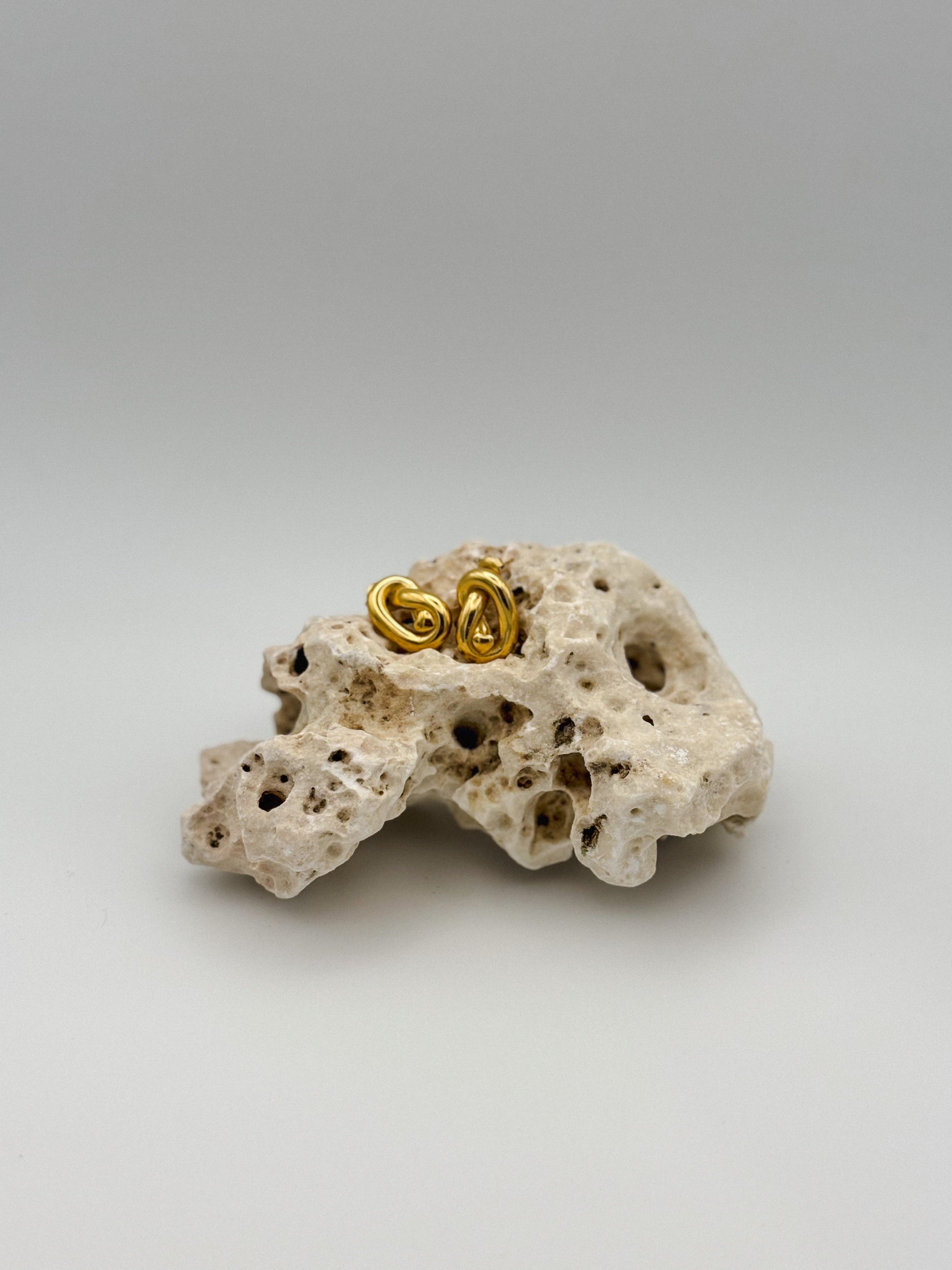 Gold abstract knot stud earrings with sculptural twist design, displayed on porous natural stone — modern artistic jewelry with a minimalist edge
