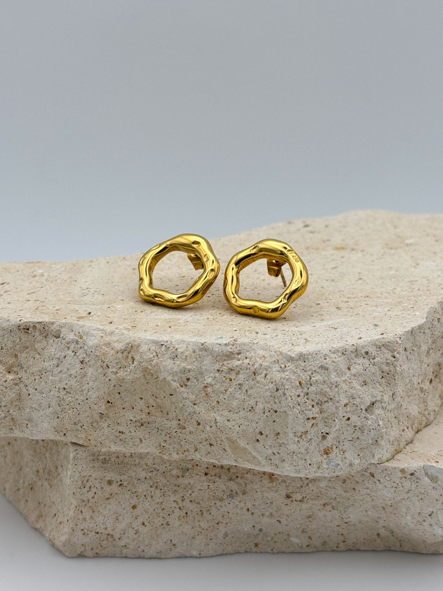 Gold open circle stud earrings with hammered organic texture, displayed on natural stone — artistic minimalist design, modern and timeless jewelry