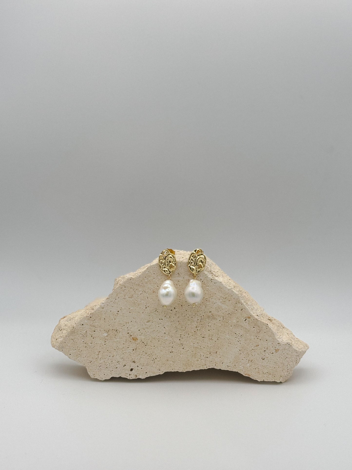 Goldene Ohrringe mit organisch geformtem Stecker und großer, barockähnlicher Perle als Anhänger, arrangiert auf hellem Naturstein.
