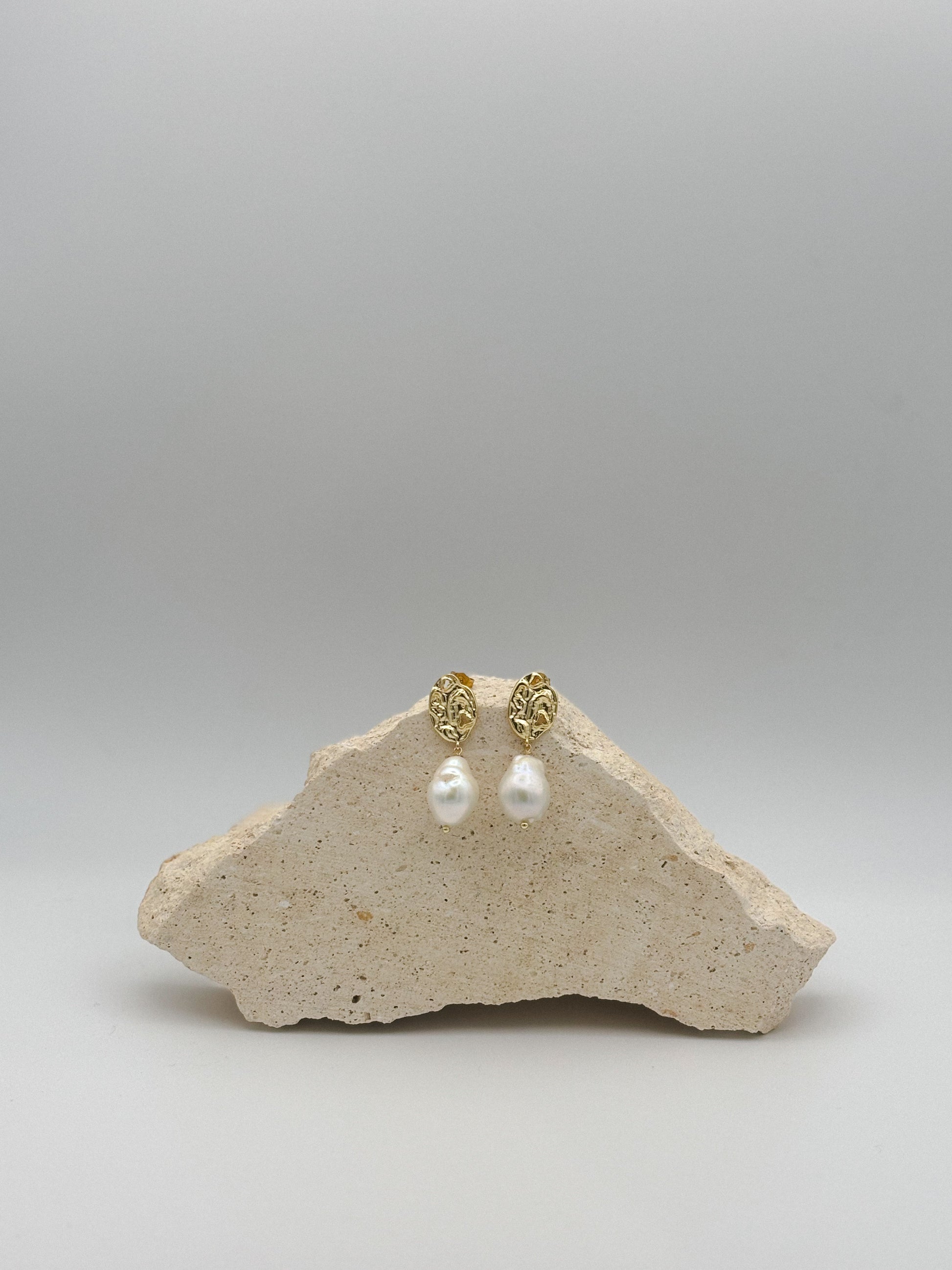 Goldene Ohrringe mit organisch geformtem Stecker und großer, barockähnlicher Perle als Anhänger, arrangiert auf hellem Naturstein.
