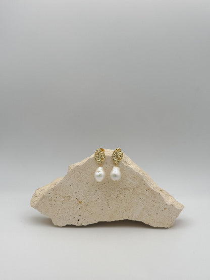 Goldene Ohrringe mit organisch geformtem Stecker und großer, barockähnlicher Perle als Anhänger, arrangiert auf hellem Naturstein.