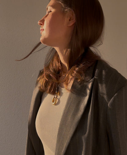 Woman wearing layered stainless steel pendant necklace on black cord, styled with neutral tank top and oversized pinstripe blazer — modern minimalist jewelry with artistic flair in warm natural light