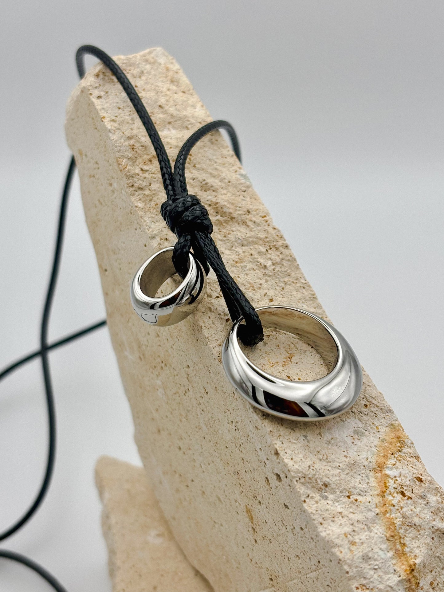 Minimalist necklace with polished silver rings on a black adjustable cord, displayed on stacked natural stone — unisex modern jewelry with clean, sculptural design