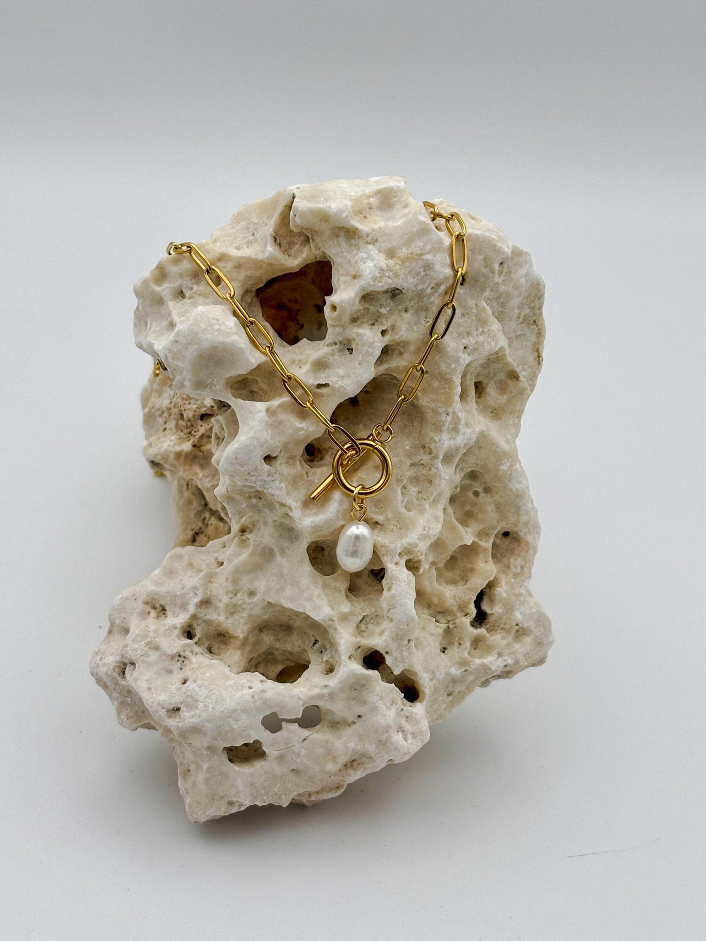 Gold paperclip chain necklace with toggle clasp and dangling freshwater pearl pendant, draped over porous natural stone — modern minimalist jewelry with organic elegance