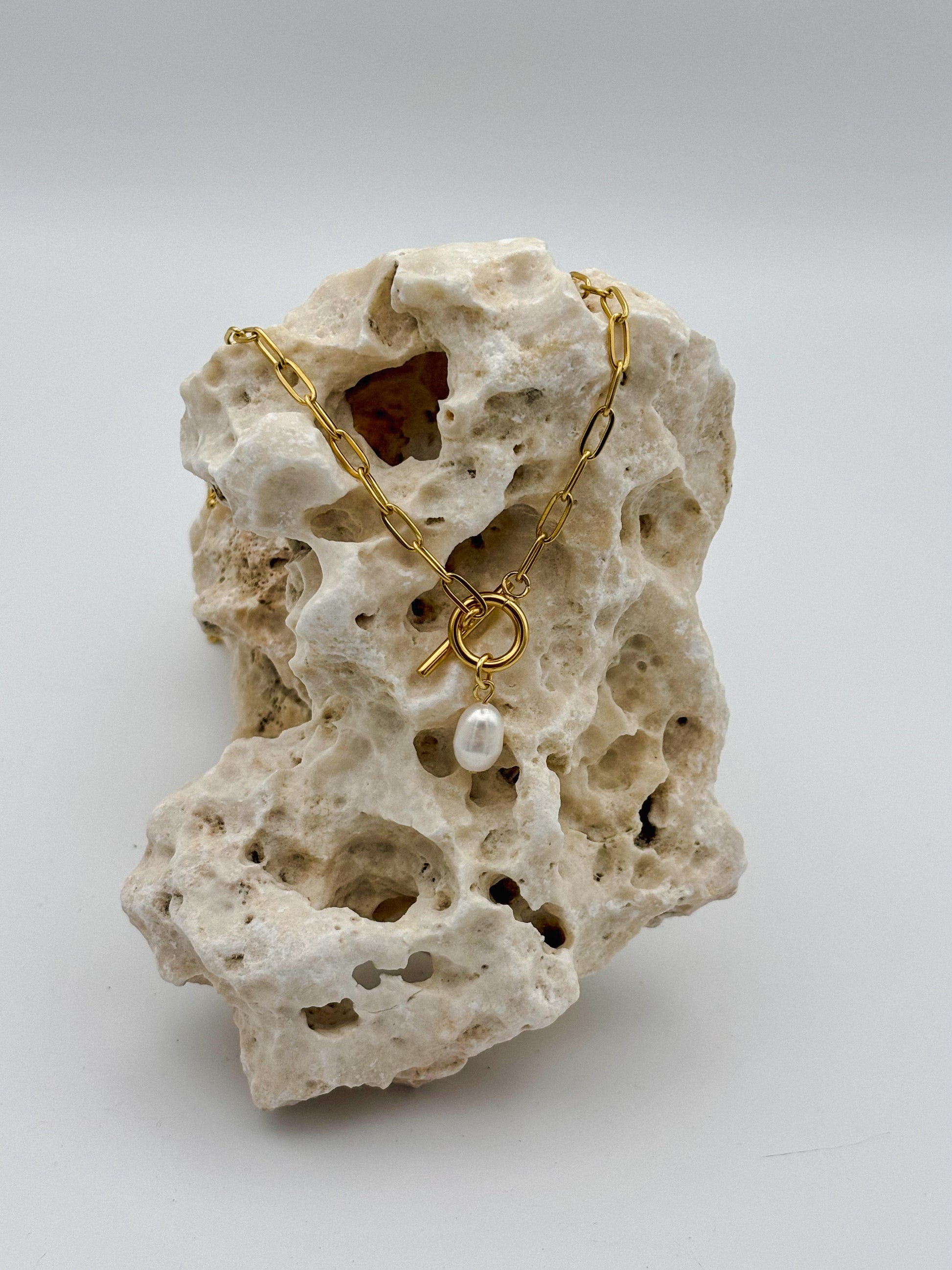 Gold paperclip chain necklace with toggle clasp and dangling freshwater pearl pendant, draped over porous natural stone — modern minimalist jewelry with organic elegance