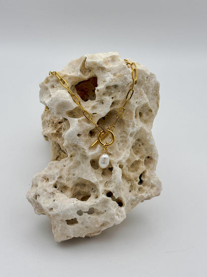 Gold paperclip chain necklace with toggle clasp and dangling freshwater pearl pendant, draped over porous natural stone — modern minimalist jewelry with organic elegance
