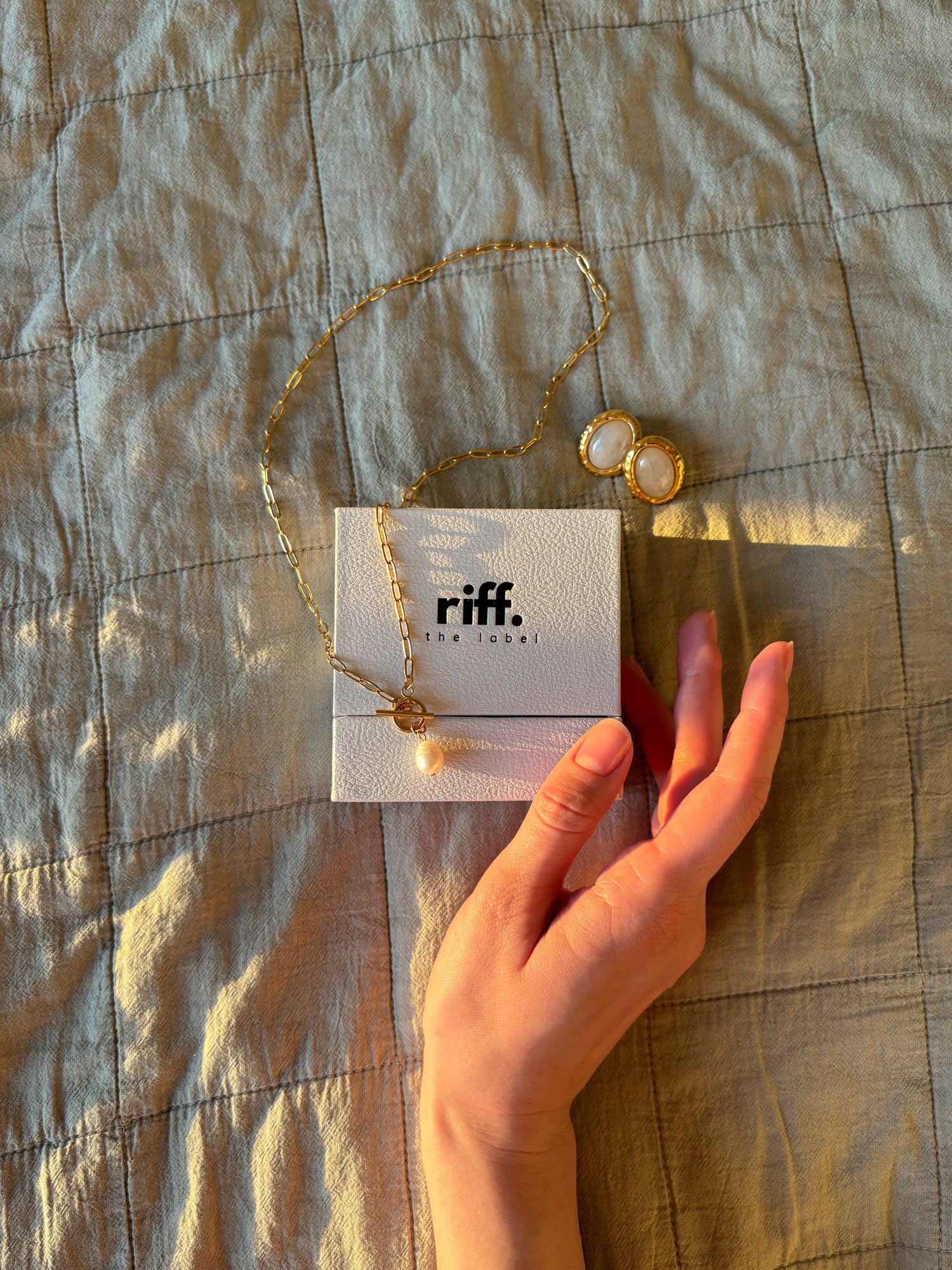 Gold chain necklace with pearl pendant from riff the label, displayed on branded jewelry box alongside matching gold and pearl earrings, hand and lemon in warm natural sunlight